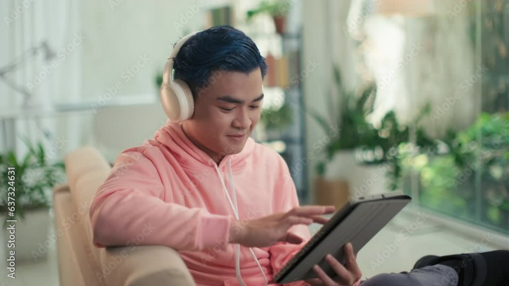 Medium shot of cheerful young man with blue hair in headphones scrolling through feed with jokes on digital tablet while resting on couch in living room Medium shot of cheerful young man with blue hair in headphones scrolling through feed with jokes on digital tablet while resting on couch in living room