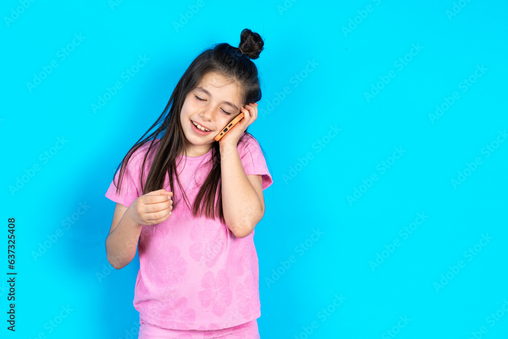 Funny Caucasian kid girl wearing pink t-shirt over blue background laughs happily, has phone conversation, being amused by friend, closes eyes. Funny Caucasian kid girl wearing pink t-shirt over blue background laughs happily, has phone conversation, being amused by friend, closes eyes.