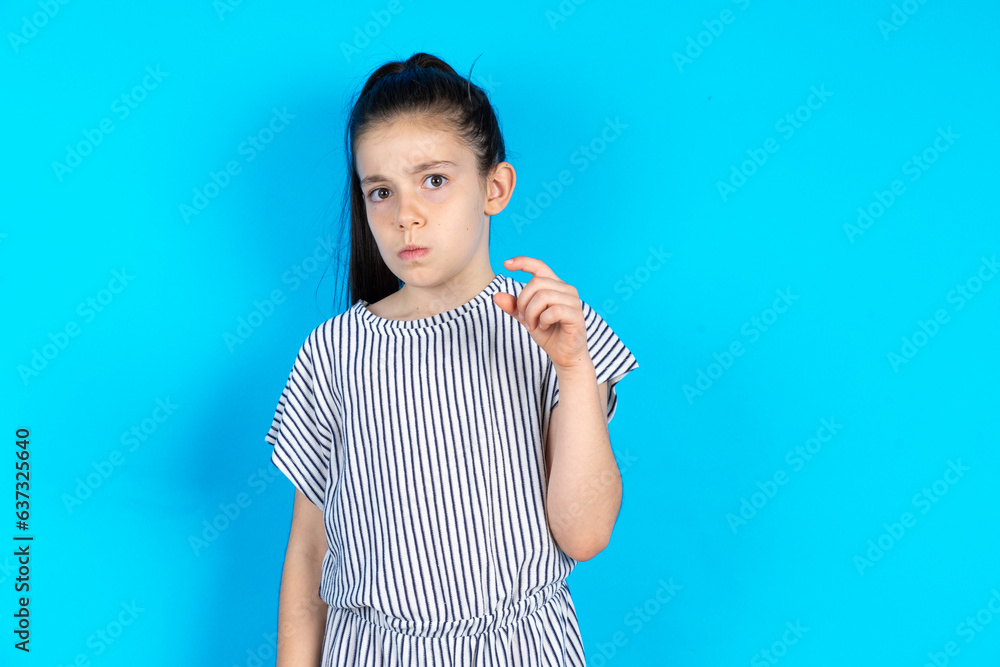 Displeased Caucasian kid girl wearing striped dress over blue background shapes little hand sign demonstrates something not very big. Body language concept. Displeased Caucasian kid girl wearing striped dress over blue background shapes little hand sign demonstrates something not very big. Body language concept.