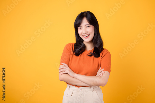 Embrace confident presence with a young Asian woman in her 30s, donning an orange shirt, showcasing crossed arm sign gesture on yellow background.