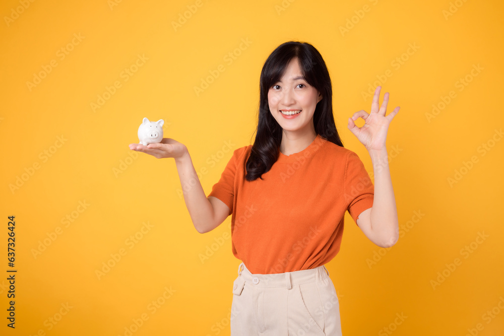 Happy Asian woman in her 30s holding piggy bank, showing okay sign, on vibrant yellow background. Wealth saving concept. Happy Asian woman in her 30s holding piggy bank, showing okay sign, on vibrant yellow background. Wealth saving concept.