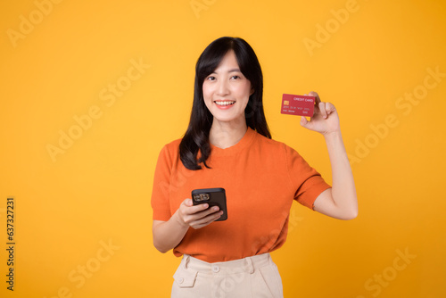 Foto Excited Asian woman 30s in orange shirt, using smartphone and showing credit card on yellow background