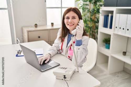 Young blonde woman doctor using laptop talking on telephone at clinic
