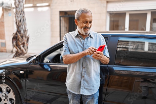 Senior grey-haired man using smartphone standing by car at street