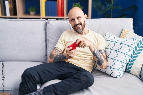 Young bald man playing video game sitting on sofa at home