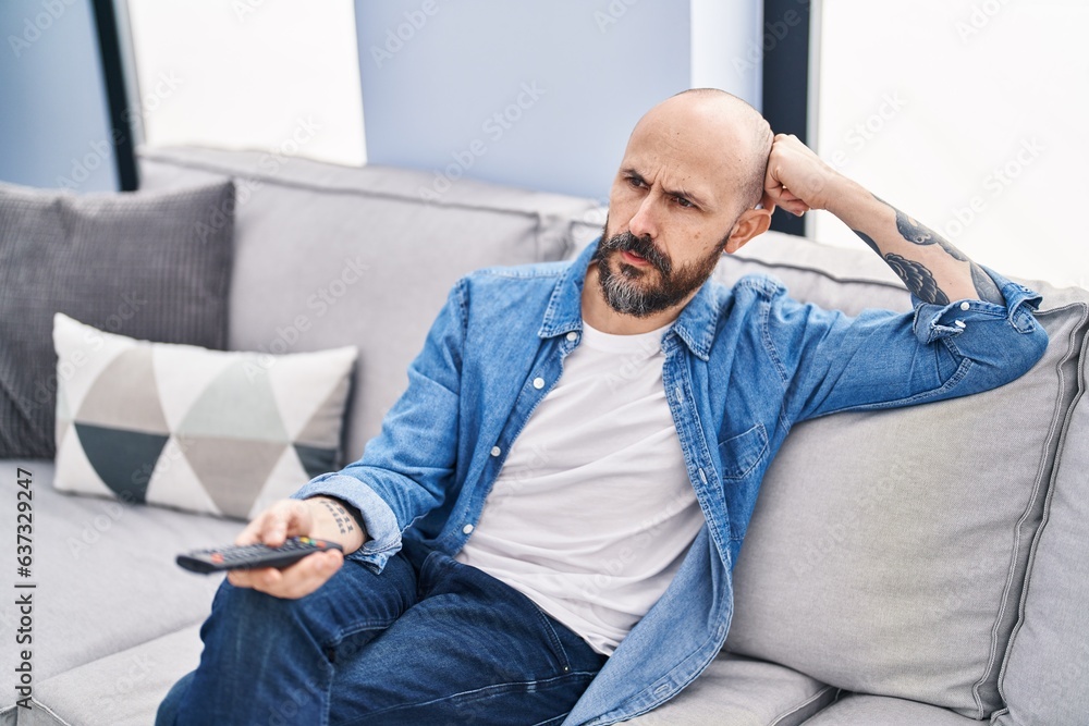 Young bald man watching tv sitting on sofa with boring expression at home Young bald man watching tv sitting on sofa with boring expression at home