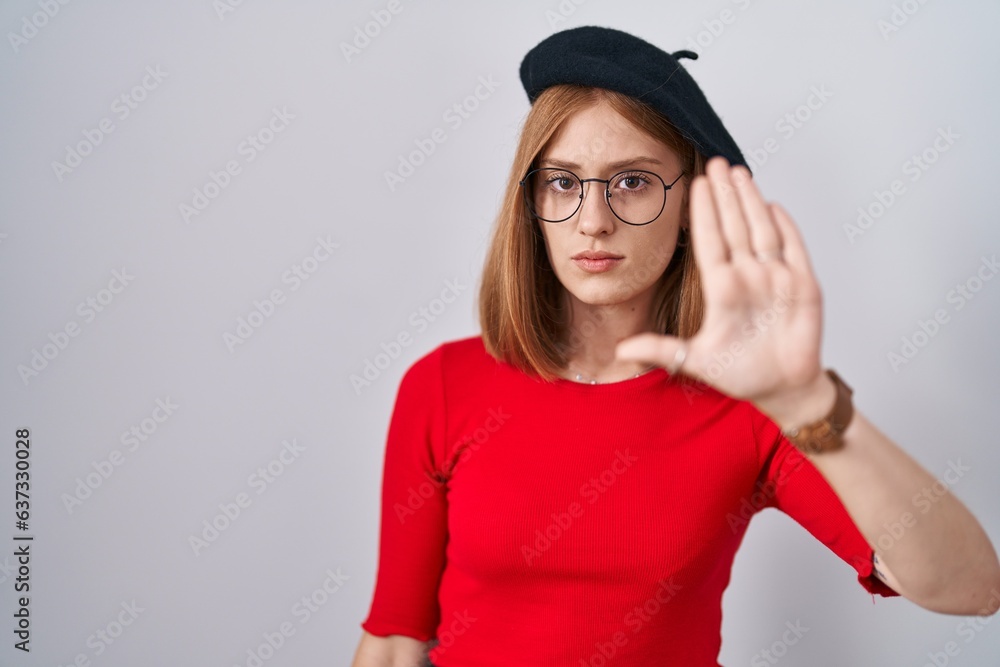 Young redhead woman standing wearing glasses and beret doing stop sing with palm of the hand. warning expression with negative and serious gesture on the face. Young redhead woman standing wearing glasses and beret doing stop sing with palm of the hand. warning expression with negative and serious gesture on the face.