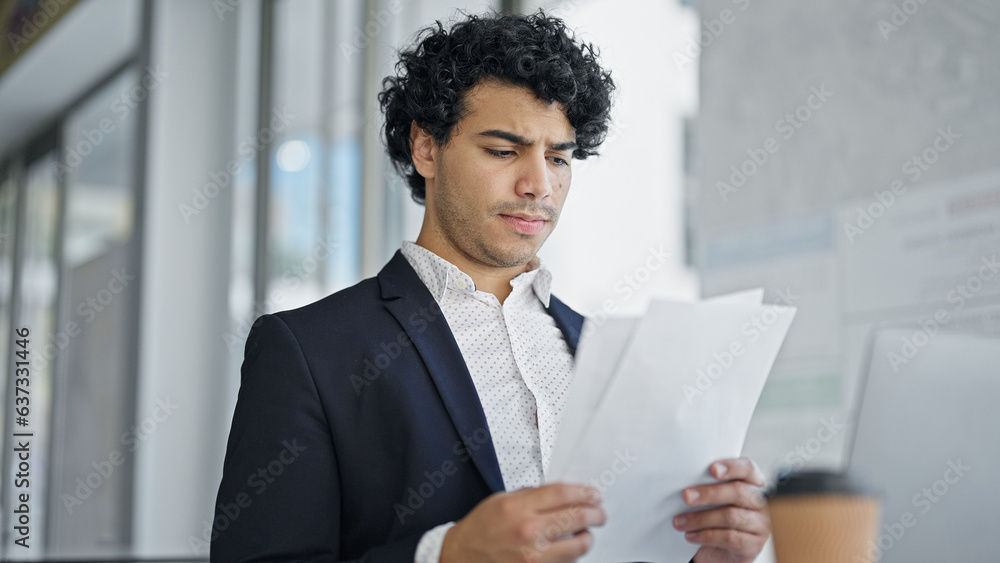 Young latin man business worker reading document sitting on table at office Young latin man business worker reading document sitting on table at office