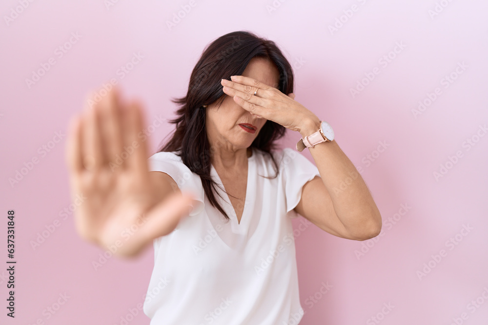 Middle age hispanic woman wearing casual white t shirt and glasses covering eyes with hands and doing stop gesture with sad and fear expression. embarrassed and negative concept. Middle age hispanic woman wearing casual white t shirt and glasses covering eyes with hands and doing stop gesture with sad and fear expression. embarrassed and negative concept.
