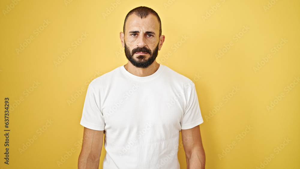 Young hispanic man standing with serious expression over isolated yellow background Young hispanic man standing with serious expression over isolated yellow background
