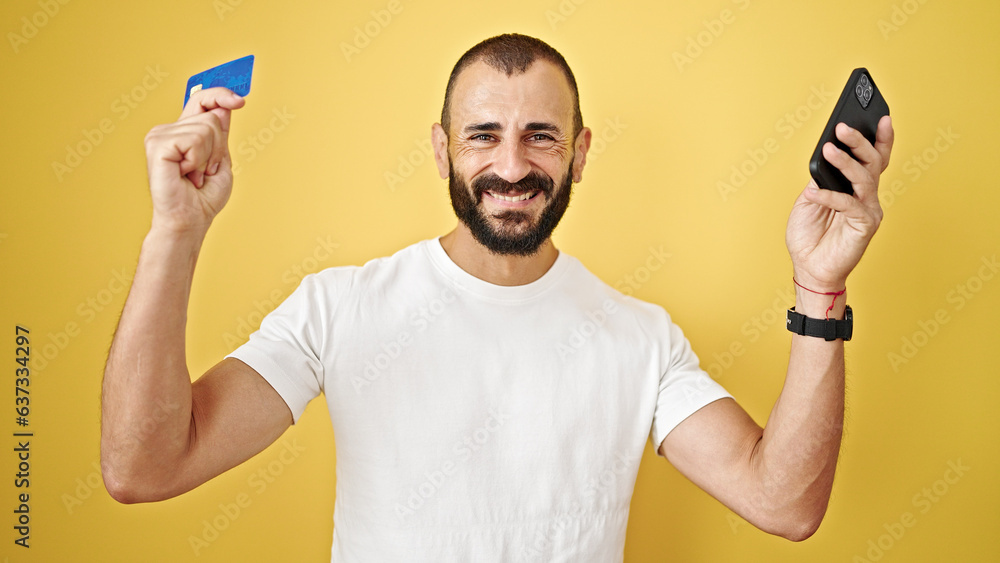Young hispanic man shopping with smartphone and credit card over isolated yellow background Young hispanic man shopping with smartphone and credit card over isolated yellow background