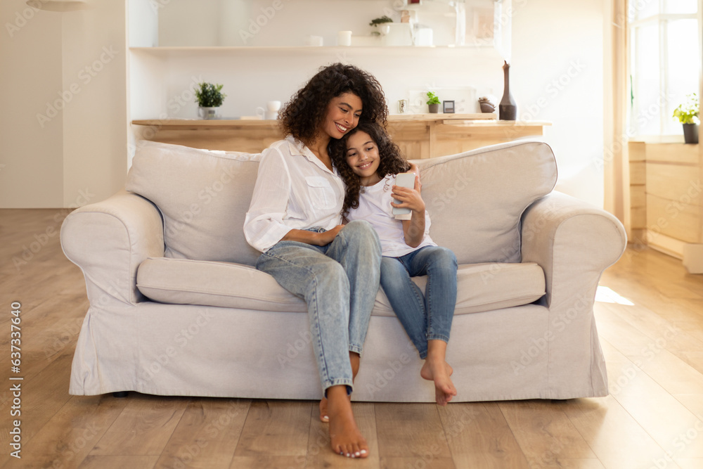 Adorable latin mother and daughter using cellphone, sitting on sofa in living room, spending time together at home, having video chat, copy space Adorable latin mother and daughter using cellphone, sitting on sofa in living room, spending time together at home, having video chat, copy space