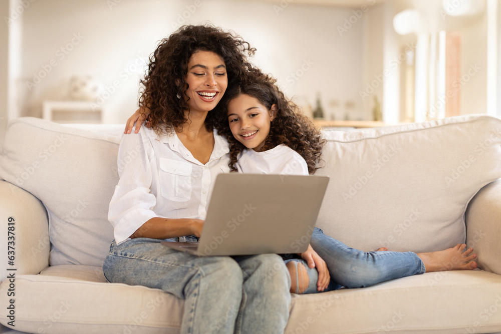 Family digital leisure. Cheerful latin mother and daughter using laptop engaging in fun online activities, surfing internet or video calling sitting on sofa at home, free space - obrazy, fototapety, plakaty 