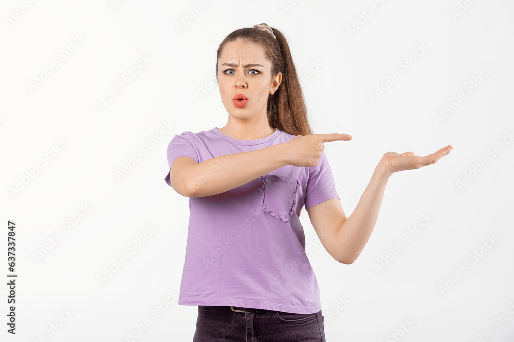 Surprised young woman with long hair points finger at banner, shows great promo offer aside, looking amazed by promotion offer, stands in casual t shirt over white background Surprised young woman with long hair points finger at banner, shows great promo offer aside, looking amazed by promotion offer, stands in casual t shirt over white background