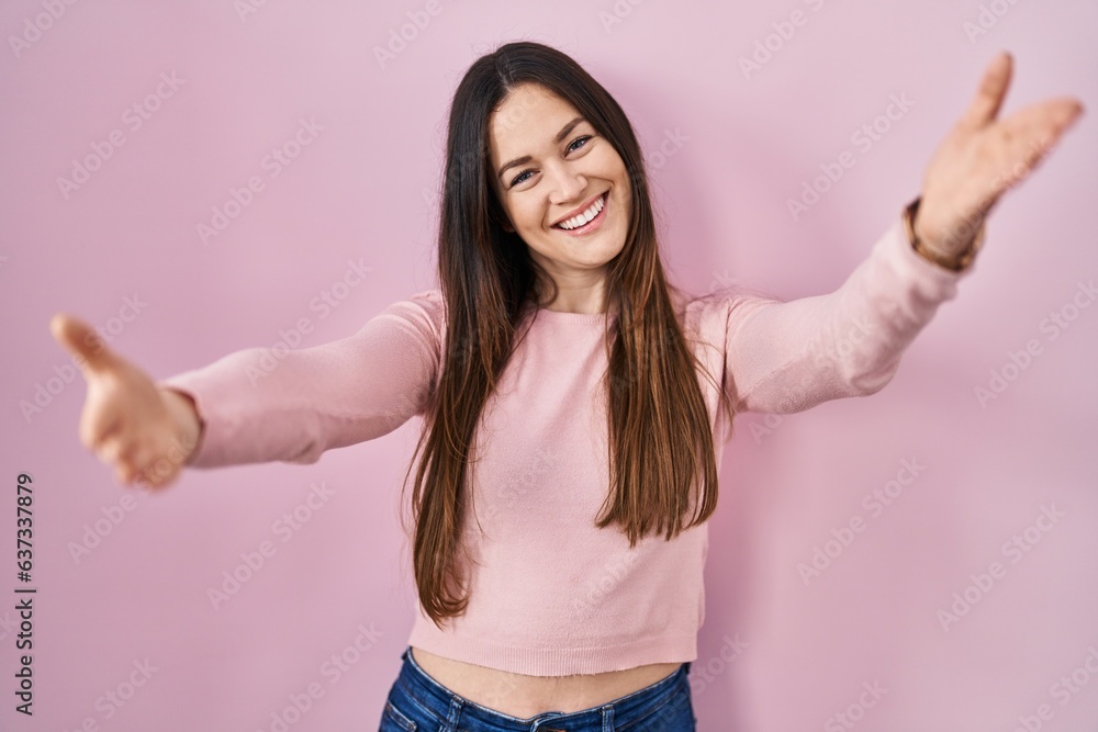 Young brunette woman standing over pink background looking at the camera smiling with open arms for hug. cheerful expression embracing happiness. Young brunette woman standing over pink background looking at the camera smiling with open arms for hug. cheerful expression embracing happiness.