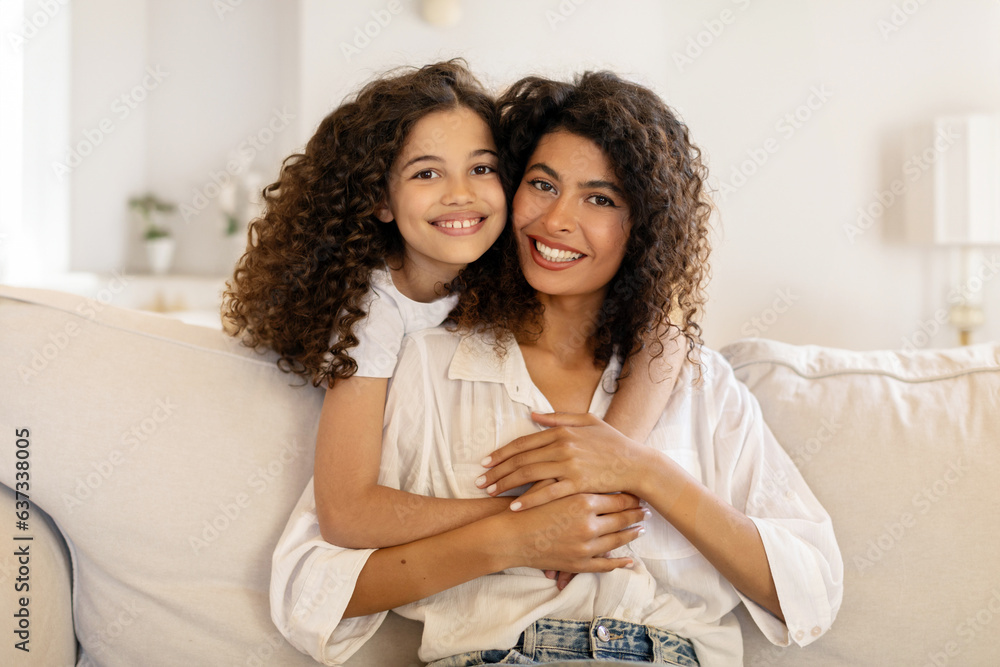 Happy latin girl hugging her beautiful mother and smiling together at camera, sitting on sofa in living room, adorable family embracing at home - obrazy, fototapety, plakaty 