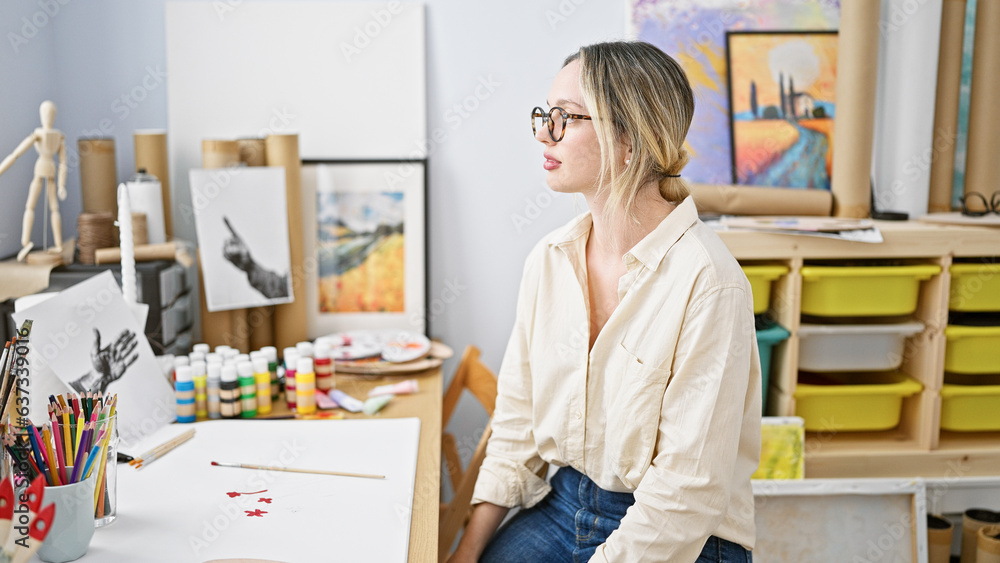 Young blonde woman artist sitting on table with serious face at art studio Young blonde woman artist sitting on table with serious face at art studio