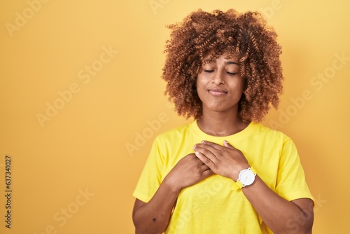 Young hispanic woman with curly hair standing over yellow background smiling with hands on chest with closed eyes and grateful gesture on face. health concept.