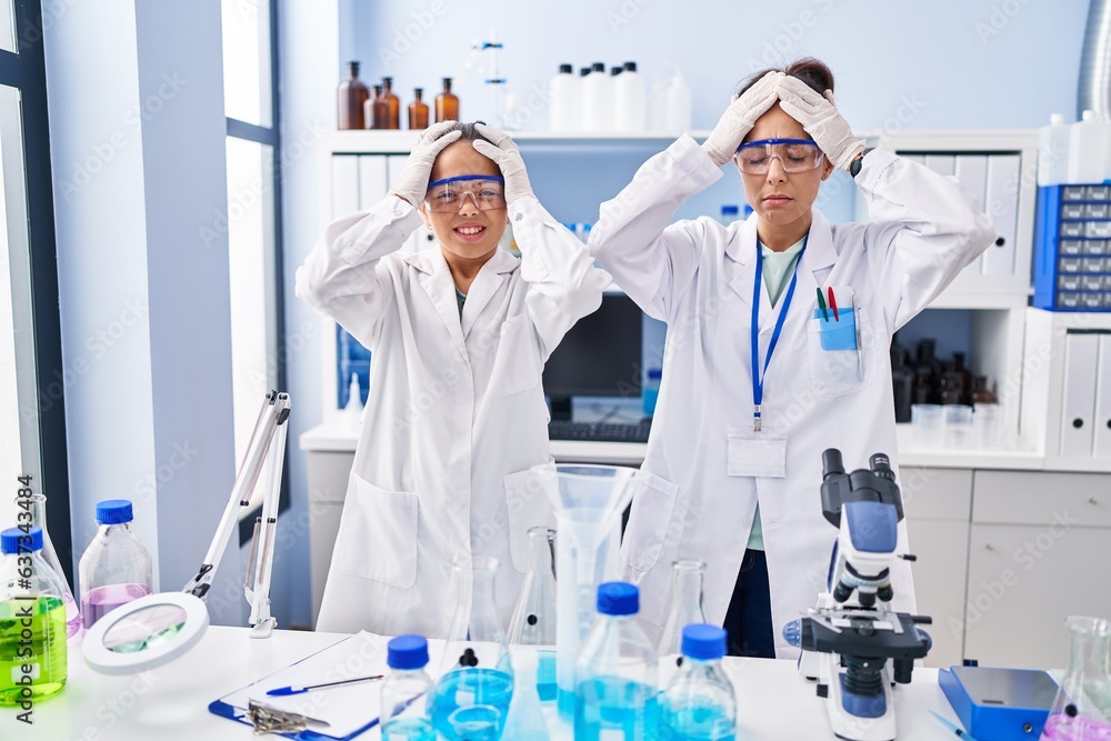 Young mother and daughter at scientist laboratory suffering from headache desperate and stressed because pain and migraine. hands on head. Young mother and daughter at scientist laboratory suffering from headache desperate and stressed because pain and migraine. hands on head.