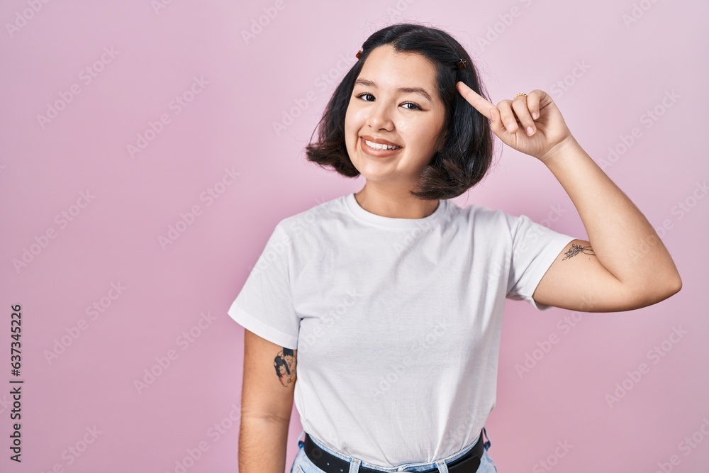 Young hispanic woman wearing casual white t shirt over pink background smiling pointing to head with one finger, great idea or thought, good memory Young hispanic woman wearing casual white t shirt over pink background smiling pointing to head with one finger, great idea or thought, good memory