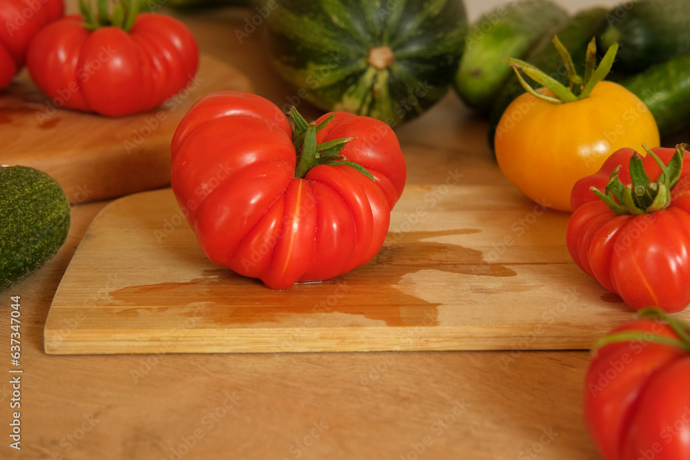 One fresh ripe big tomato on a wooden cutting board. Many raw vegetables background. Tomatoes, cucumbers. Cooker at home making a healthy veggie meal. Chopping ingredient for preparing vegetarian dish One fresh ripe big tomato on a wooden cutting board. Many raw vegetables background. Tomatoes, cucumbers. Cooker at home making a healthy veggie meal. Chopping ingredient for preparing vegetarian dish