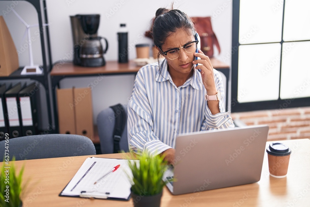 Young beautiful hispanic woman business worker using laptop talking on smartphone at office Young beautiful hispanic woman business worker using laptop talking on smartphone at office