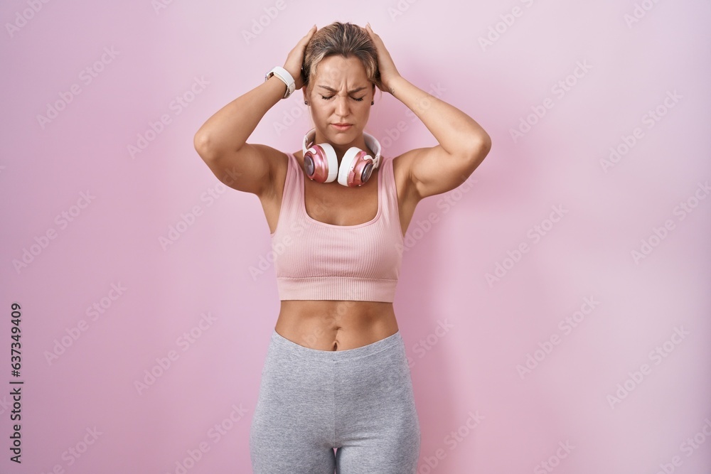 Young blonde woman wearing sportswear and headphones suffering from headache desperate and stressed because pain and migraine. hands on head. Young blonde woman wearing sportswear and headphones suffering from headache desperate and stressed because pain and migraine. hands on head.