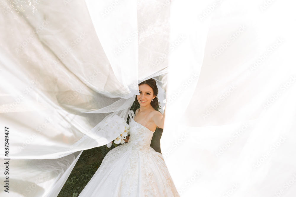 Warm sunny portrait of a happy bride with a bouquet of orchids in her hands, outdoors and at sunset. Warm summer time. A long veil, a luxurious white dress with a train. Warm sunny portrait of a happy bride with a bouquet of orchids in her hands, outdoors and at sunset. Warm summer time. A long veil, a luxurious white dress with a train.