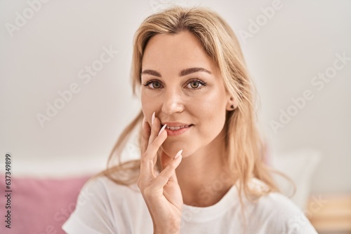 Young blonde woman smiling confident touching face at bedroom