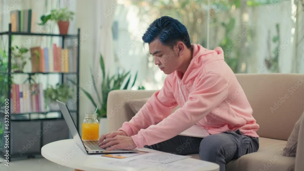 Medium long shot of serious young graphic designer with blue hair working on laptop while sitting on couch at home, fresh drink on table Medium long shot of serious young graphic designer with blue hair working on laptop while sitting on couch at home, fresh drink on table