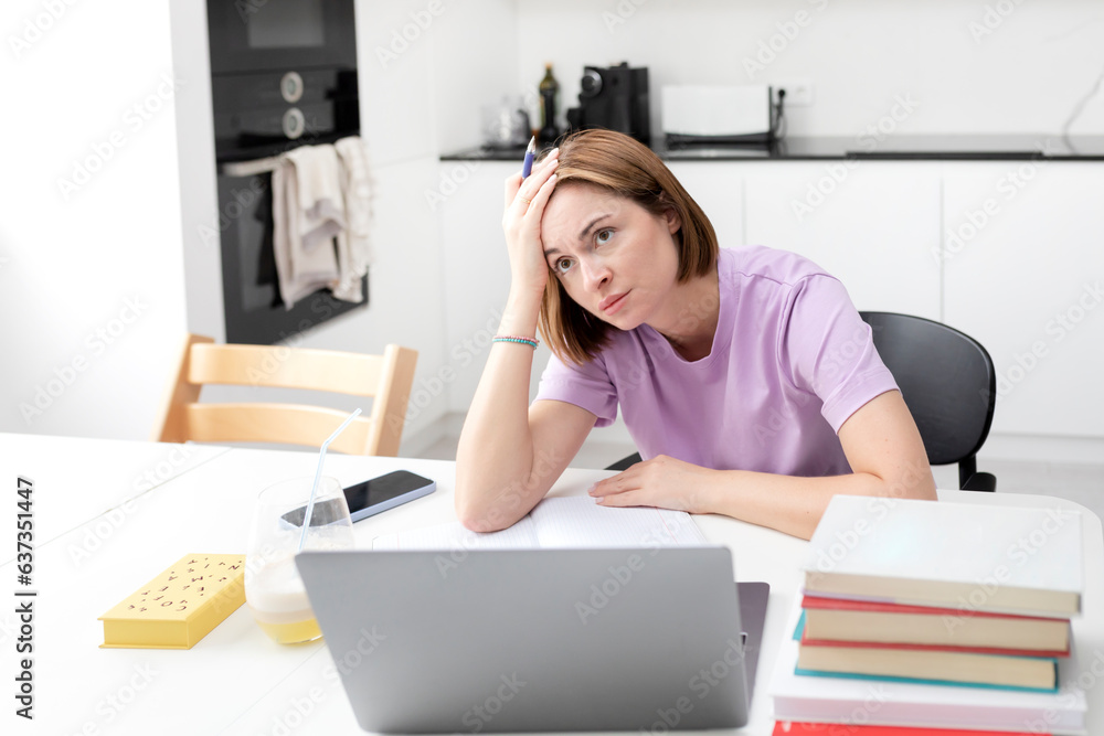 Obraz premium Tired and bored female student reading book and preparing for exams at university. Stack of books on the desk. Online education and learning from home concept
