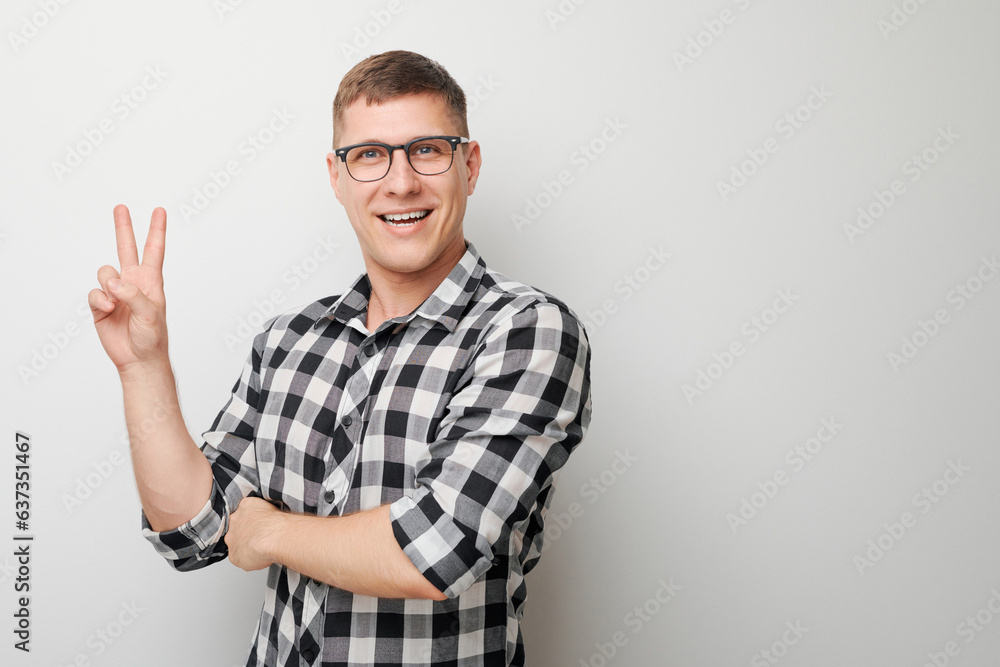 Naklejka premium Portrait of young happy caucasian man in shirt showing peace and love gesture with fingers isolated on white studio background