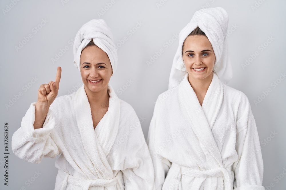 Middle age woman and daughter wearing white bathrobe and towel showing and pointing up with finger number one while smiling confident and happy.