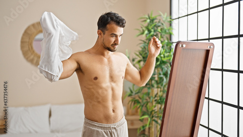 Young hispanic man taking t shirt off looking body on mirror at bedroom