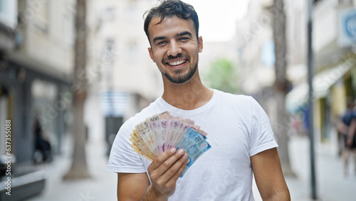 Young hispanic man smiling confident holding mexican pesos at street photo