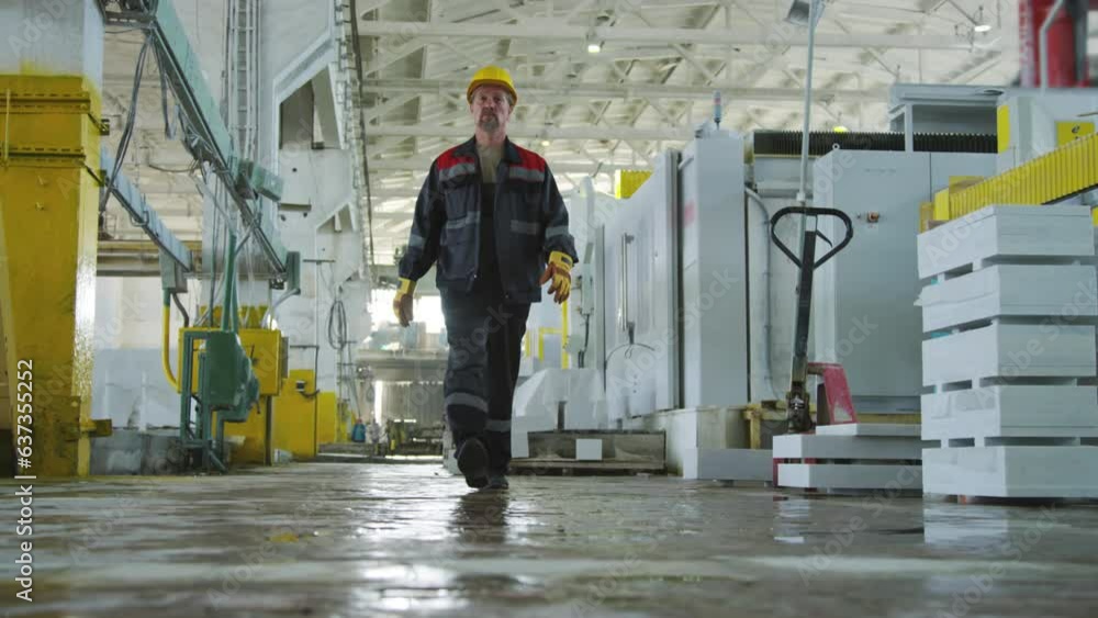 Ground level shot of middle aged man in workwear, hardhat and protective gloves walking through warehouse of stone factory during day Ground level shot of middle aged man in workwear, hardhat and protective gloves walking through warehouse of stone factory during day