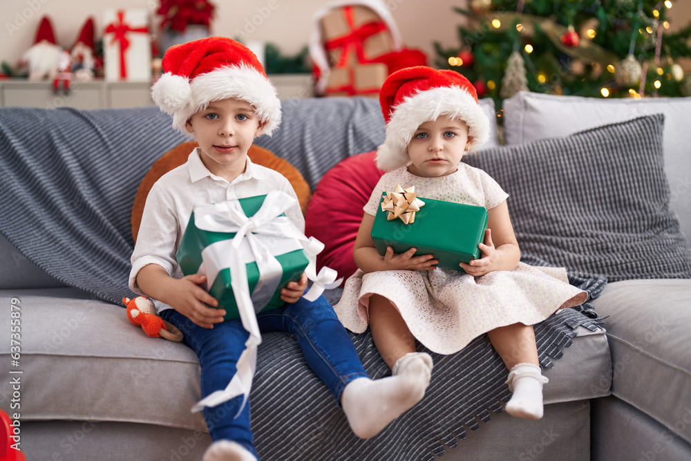 Two kids holding gifts sitting on sofa by christmas tree at home Two kids holding gifts sitting on sofa by christmas tree at home