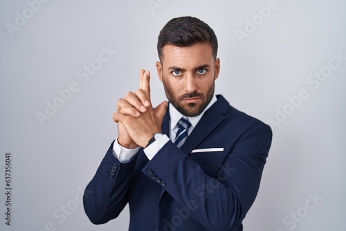 Handsome hispanic man wearing suit and tie holding symbolic gun with hand gesture, playing killing shooting weapons, angry face