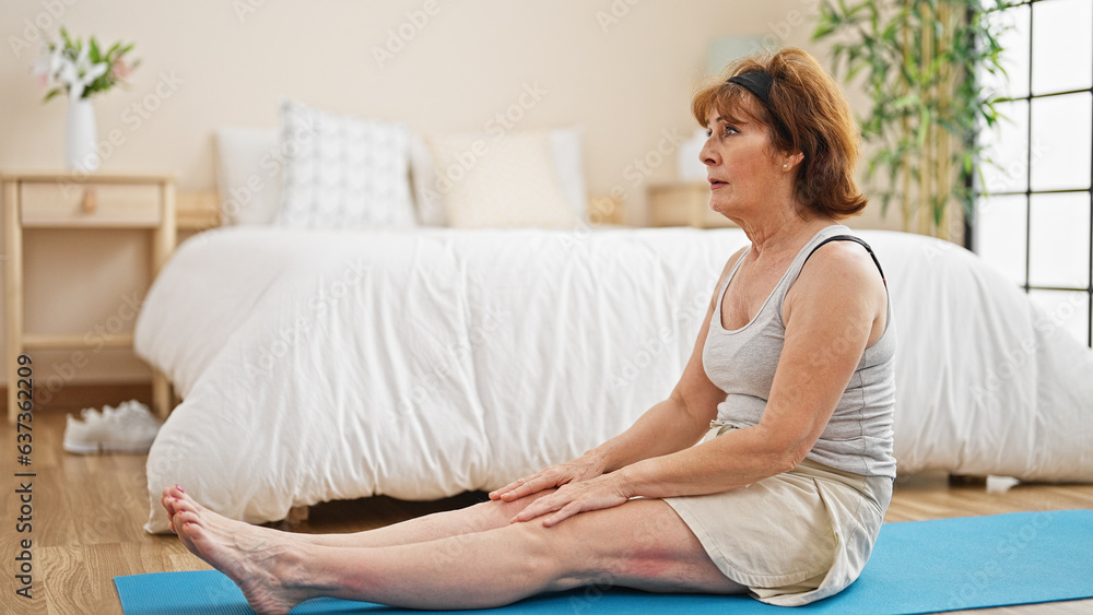 Middle age woman stretching legs sitting on yoga mat at bedroom