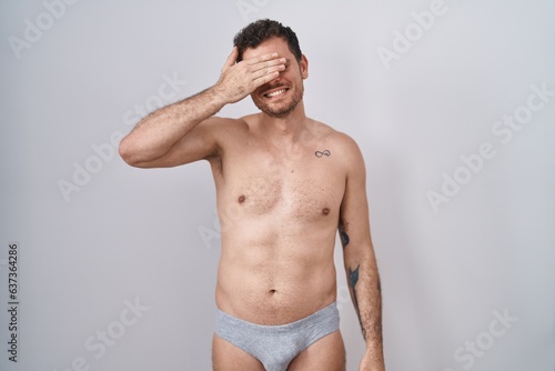 Young hispanic man standing shirtless wearing underware smiling and laughing with hand on face covering eyes for surprise. blind concept.