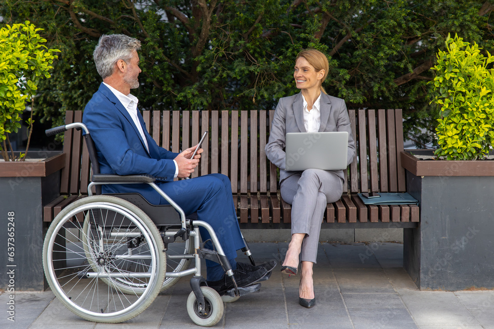 Businessman in wheelchair with female partner using laptop and tablet for working on joint project Businessman in wheelchair with female partner using laptop and tablet for working on joint project