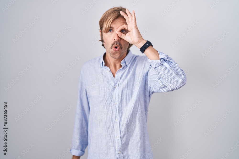 Caucasian man with mustache standing over white background doing ok gesture shocked with surprised face, eye looking through fingers. unbelieving expression. Caucasian man with mustache standing over white background doing ok gesture shocked with surprised face, eye looking through fingers. unbelieving expression.