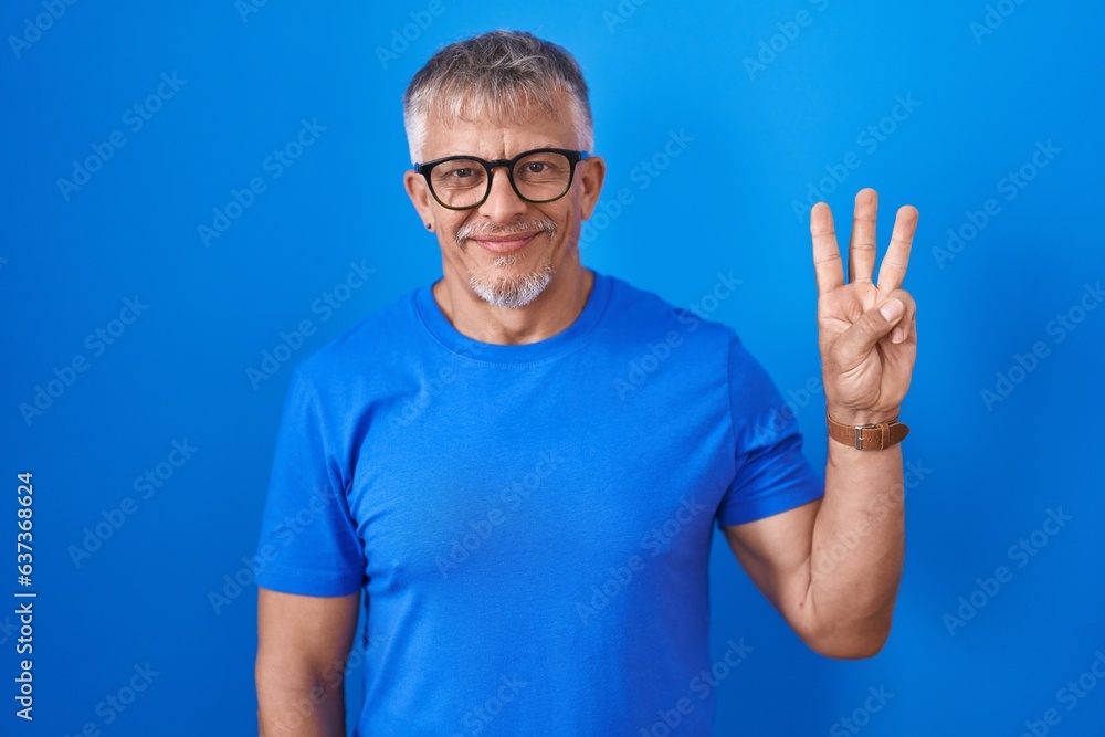 Hispanic man with grey hair standing over blue background showing and pointing up with fingers number three while smiling confident and happy. Hispanic man with grey hair standing over blue background showing and pointing up with fingers number three while smiling confident and happy.