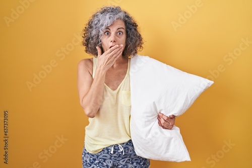 Middle age woman with grey hair wearing pijama hugging pillow shocked covering mouth with hands for mistake. secret concept.