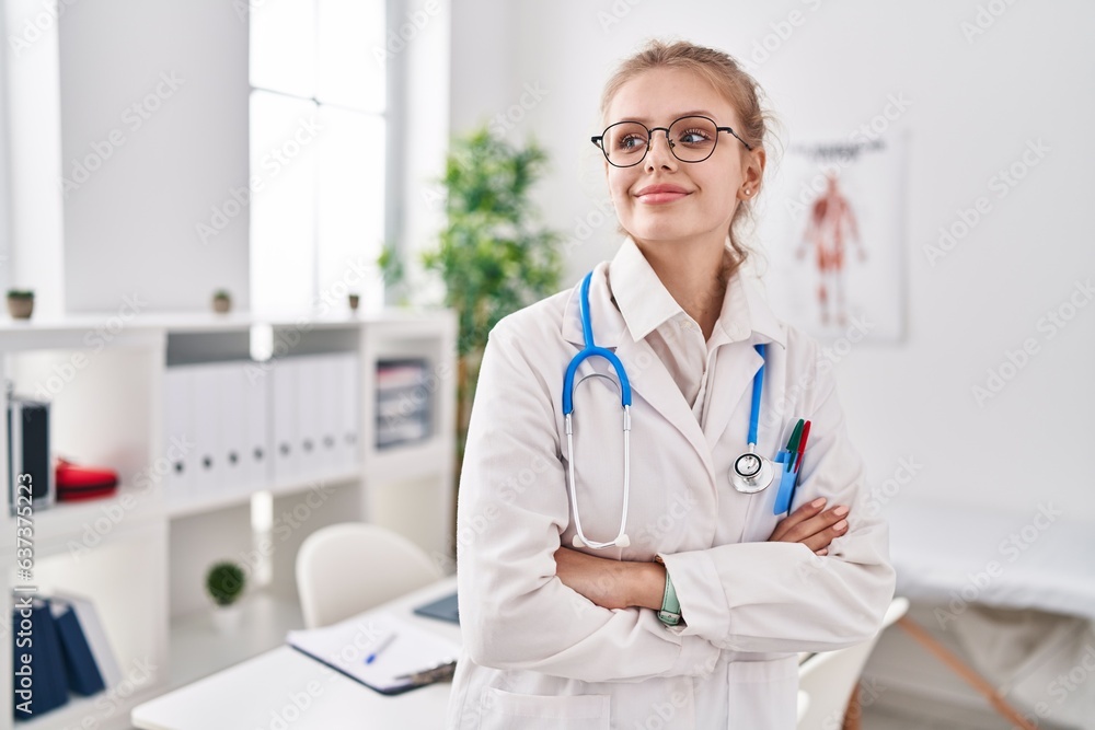 Young blonde woman doctor smiling confident standing with arms crossed gesture at clinic Young blonde woman doctor smiling confident standing with arms crossed gesture at clinic