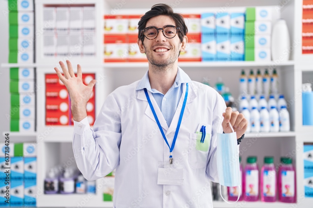 Young hispanic man working at pharmacy drugstore holding safety mask celebrating victory with happy smile and winner expression with raised hands