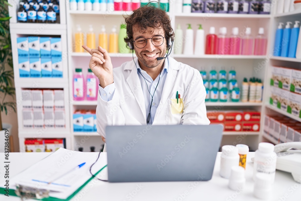 Hispanic young man working at pharmacy drugstore working with laptop smiling happy pointing with hand and finger to the side