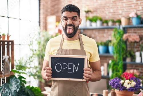 Hispanic man with beard working at florist holding open sign sticking tongue out happy with funny expression.