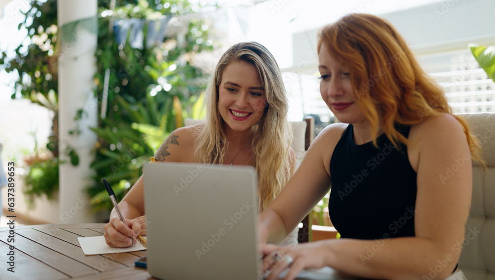 Two women using laptop writing on notebook sitting on table at home terrace