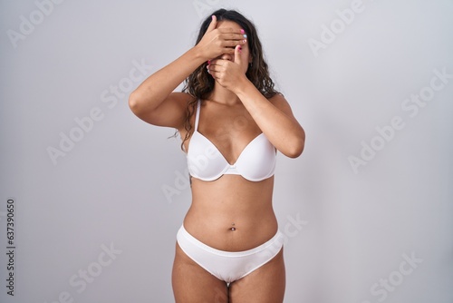 Young hispanic woman wearing white lingerie covering eyes and mouth with hands, surprised and shocked. hiding emotion © Krakenimages.com